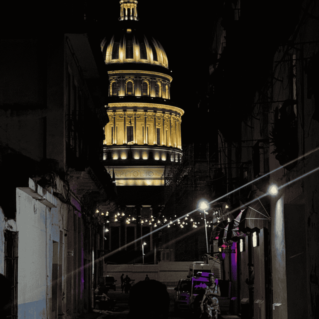 Una strada scarsamente illuminata incornicia il Campidoglio dell'Avana, Cuba illuminato di notte. Fili di luci aleggiano sopra la strada, creando un'atmosfera serena e drammatica.