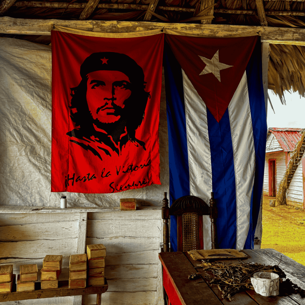 Uno scorcio di Viñales con una bandiera rossa di Che Guevara con la scritta "¡Hasta la Victoria Siempre!" e una bandiera cubana. Scatole di legno sono impilate su un mobile bianco, evocando un'atmosfera storica e rivoluzionaria.
