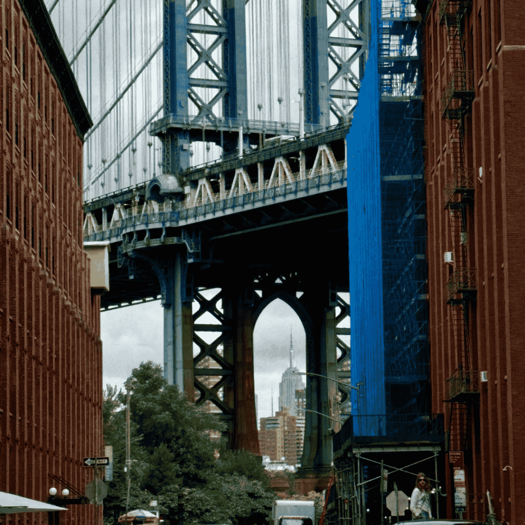 Vista del Manhattan Bridge incorniciato da edifici in mattoni rossi, con l'Empire State Building visibile in lontananza. Cielo coperto e contesto urbano. Si puo' visitare New York con un visto USA B&/B2 dopo un viaggio a Cuba.