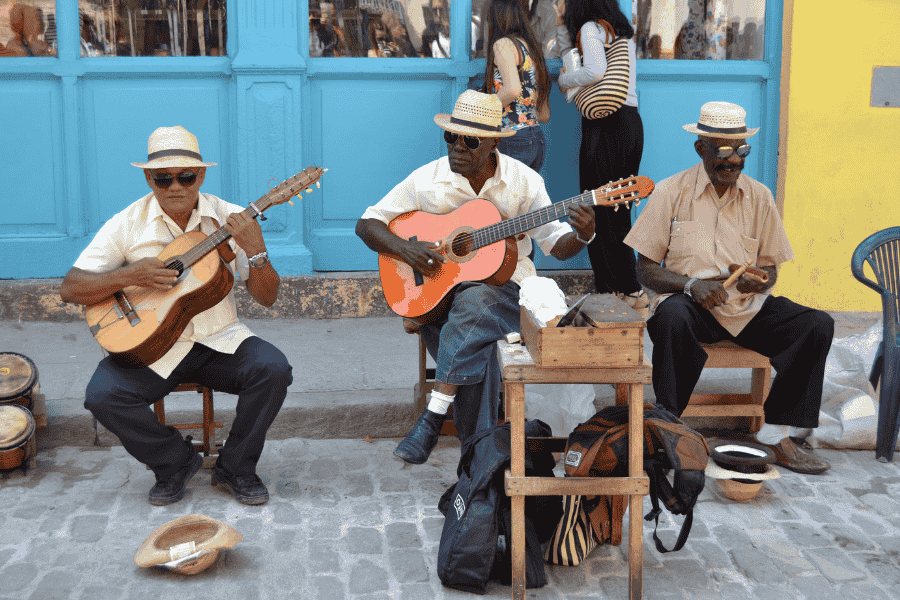 Tre musicisti di strada all'Havana, Cuba con cappelli di paglia suonano chitarre e maracas fuori da un edificio dipinto di colori vivaci. Creano un'atmosfera vivace e allegra di salsa e suoni esotici. Si puo' andare a Cuba e visitare gli Stati Uniti facendo richiesta del visto USA dopo un viaggio a Cuba.
