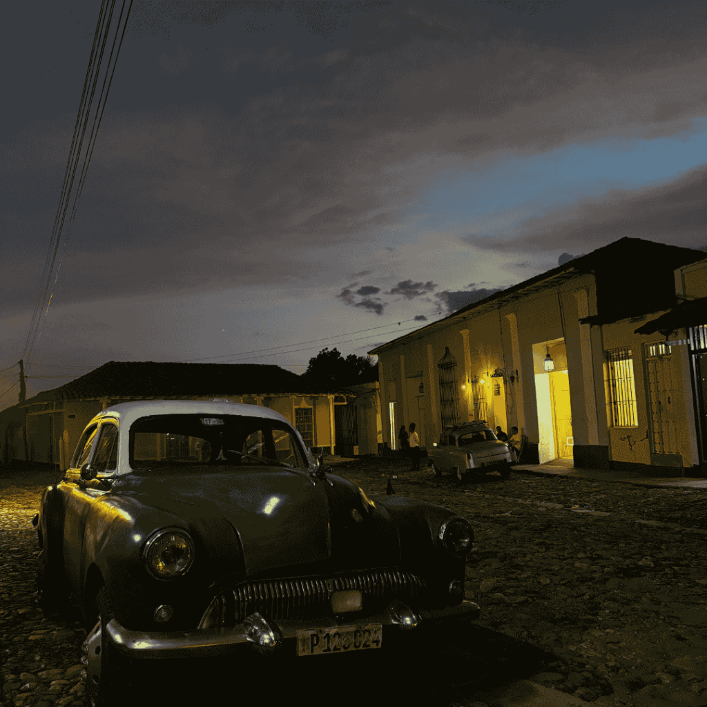 Auto d'epoca parcheggiata su una strada acciottolata di Trinidad, Cuba al tramonto, con cieli crepuscolari colorati. Edifici coloniali illuminati di giallo creano un'atmosfera nostalgica e tranquilla.