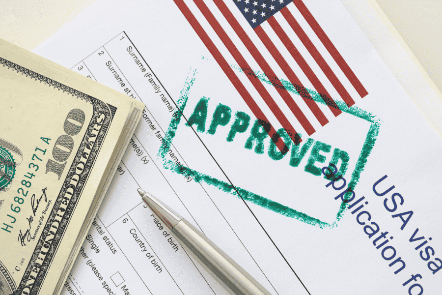 Visto USA B1/B2 dopo un viaggio a Cuba. Una pila di banconote da cento dollari USA è appoggiata su un modulo di richiesta di visto USA timbrato con "APPROVATO" in verde. Sono visibili una penna e una bandiera USA, a simboleggiare il successo e l'approvazione ufficiale.
